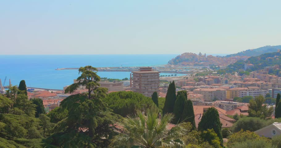 Elevated panoramic view of Imperia, Italy, featuring Mediterranean architecture, terracotta rooftops, lush green trees, a coastal harbor and the blue Ligurian Sea.