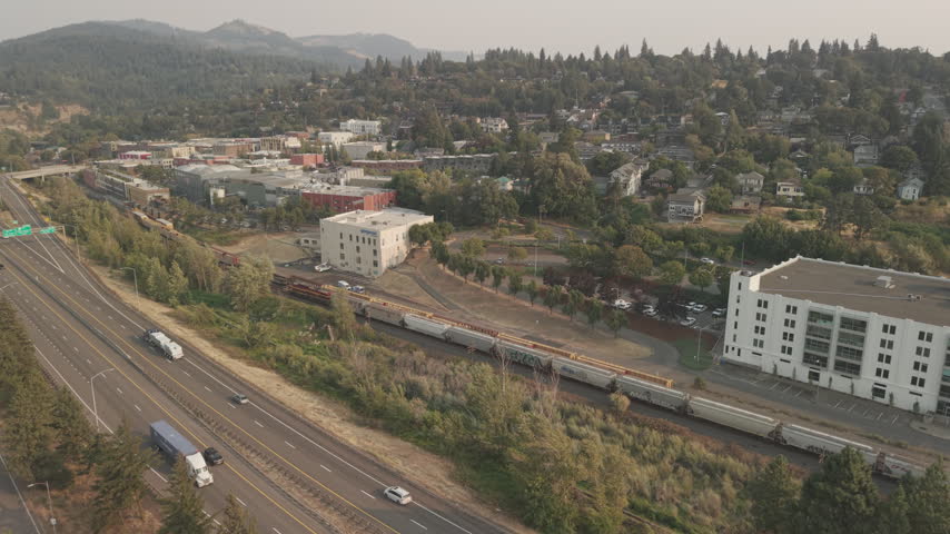 Freight train along Interstate 84 in Hood River, Oregon, with residential neighborhoods, trees, and hazy summer conditions in the Columbia River Gorge.