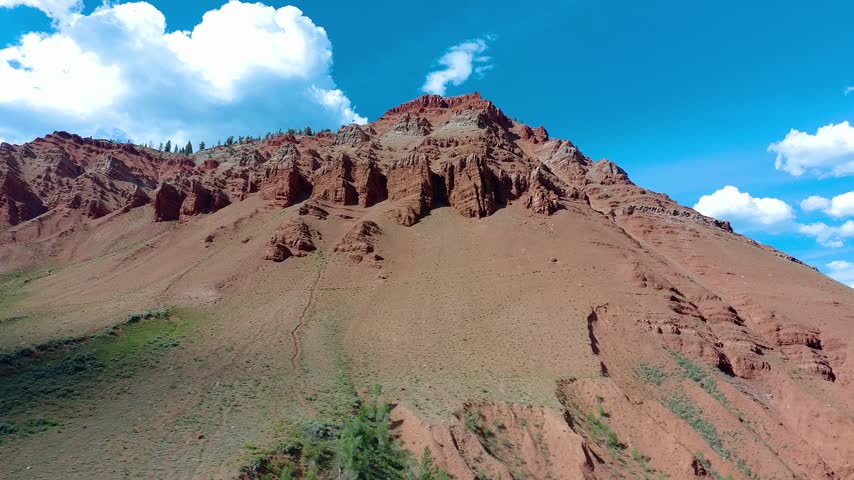 Red rock mountain slopes under bright blue sky. White clouds drift above rugged terrain. Green shrubs cling to rocky foothills. Vibrant earth tones dominate the landscape