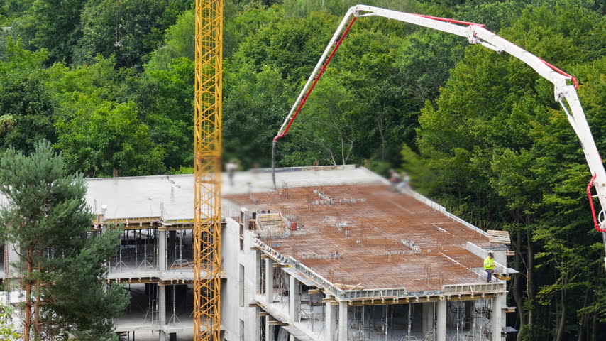 A concrete pump truck boom is positioned over a multi-story building roof during a technical break in construction. A yellow tower crane stands high next to the modern reinforced structure.