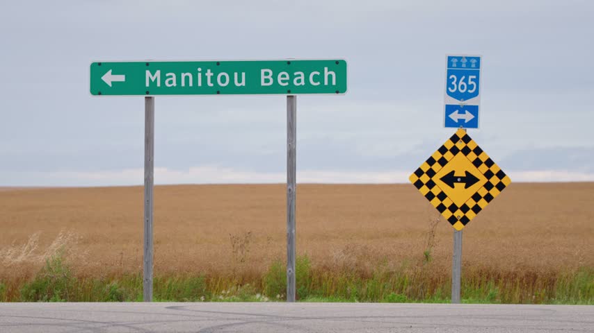 Static close-up of the Manitou Beach roadside sign pointing left, with a road in front and a uniform golden wheat field behind in rural Saskatchewan, Canada