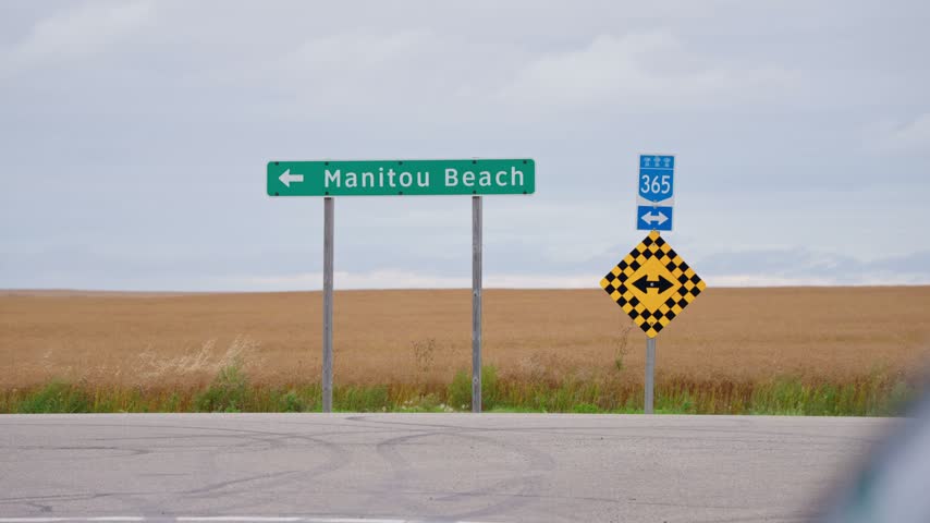 Static wider shot of the Manitou Beach roadside sign at a rural Saskatchewan intersection, with a road in front and a golden wheat field stretching behind the sign