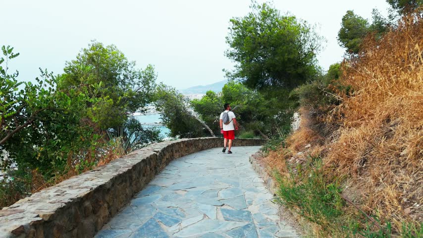 A man with a backpack walks along the Alcazaba path surrounded by greenery and flowers, with glimpses of the Malaga coastline beyond.