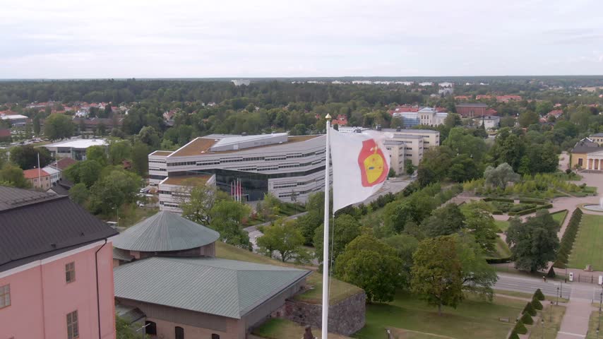 Drone by pink castle wall with Uppsala coat-of-arms flag; green roofs and campus buildings beyond trees.