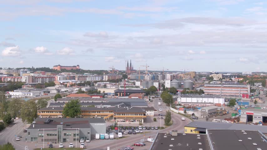 High drone past warehouses and parking toward Uppsala skyline with twin cathedral spires and pink castle.