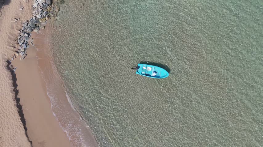Aerial view of a small blue boat floating in clear shallow water near a sandy beach in Famagusta, Famagusta, Northern Cyprus.