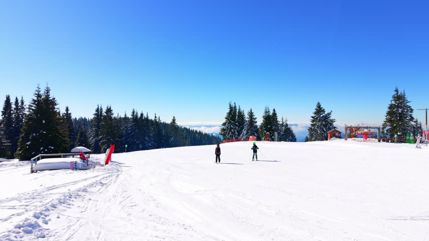 People skiing on mountain summit plateau. A clear blue sky, evergreen trees and a red chairlift station in the background.