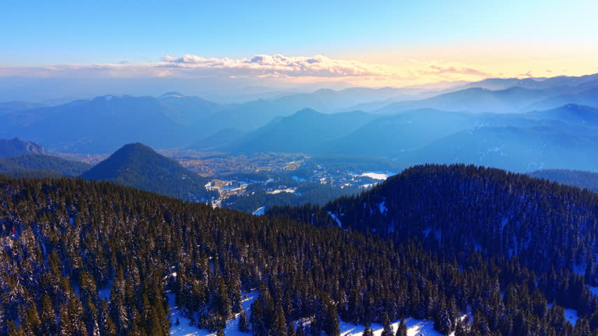 Golden sunset over layered Rhodope mountain ridges, Bulgaria. Forested slopes descend toward a distant valley town partly obscured by low hanging cloud.