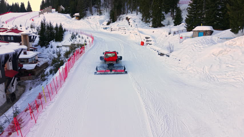 Corduroy pattern on groomed piste beside hotels. A red snow groomer drives along the slope, fenced by netting, near chalet style buildings and spruce trees.