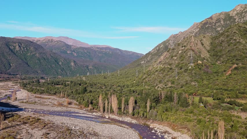 Panoramic view of a rocky riverbed bordered by a row of poplar trees, dense forested hillsides and distant high tension towers beneath the peaks