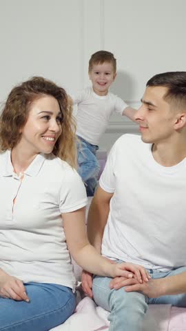 Happy family embraces on sofa as young son stands between his parents. Smiling mother and father share tender moment with their child in bright, comfortable living room, hugging their son close.