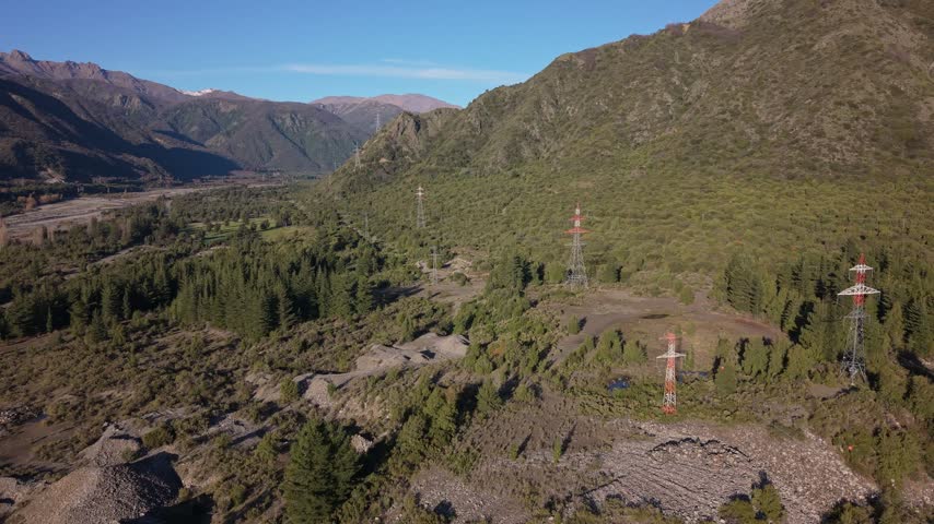 Wide aerial view of red and white high tension towers scattered across forested hillsides beside a rocky riverbed beneath rugged mountains