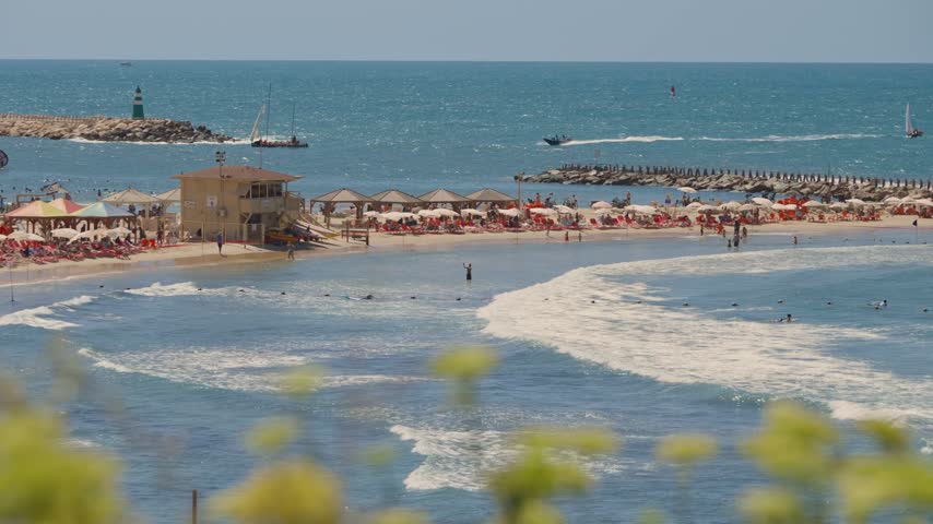 Scenic summer view of a beach in Tel Aviv, Israel, with tourists swimming, surfing, and relaxing under umbrellas while boats sail on the beautiful Mediterranean Sea on a sunny day