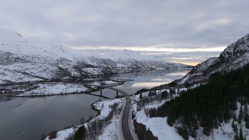 Aerial view of the Lofoten landscape featuring a bridge spanning across a calm fjord surrounded by snow-covered mountains and pine forests in Vagan Municipality, Nordland, Norway.