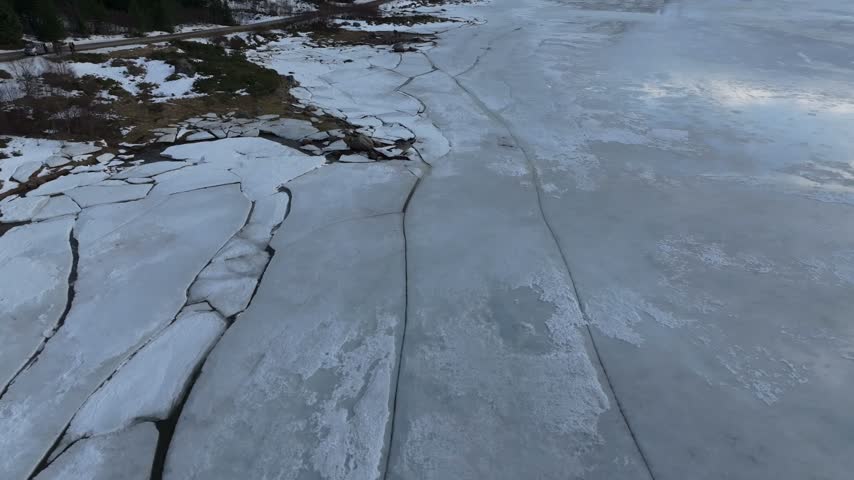 Aerial view of cracked ice on a frozen lake with a snowy shoreline and a road in the distance in Vagan Municipality, Nordland, Norway.