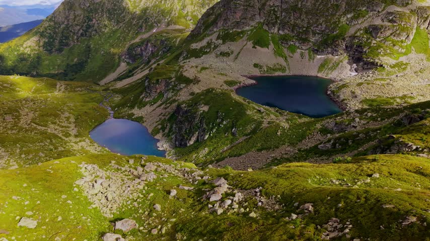 An aerial view shows forward movement over a rocky ridge toward alpine lakes in a mountain valley. The landscape features steep rocky slopes and green terrain under natural daylight.