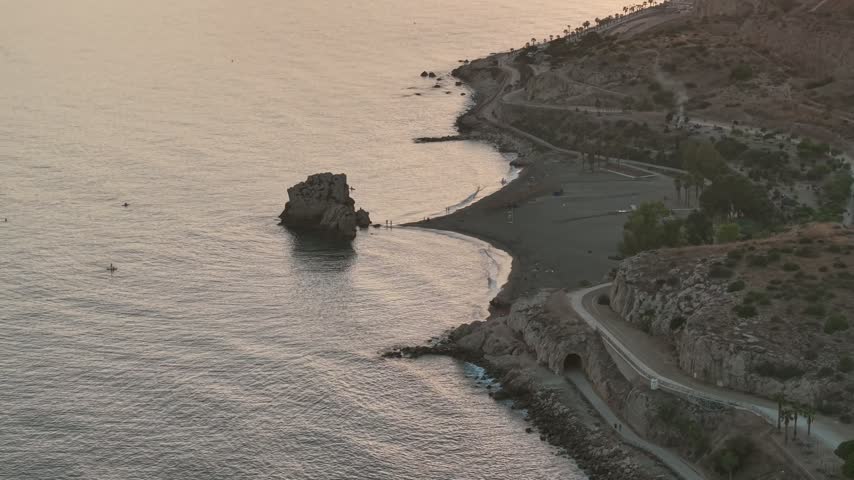 Drone view of the Peñón del Cuervo rock off a dark sand beach east of Malaga, with a coastal path, an old tunnel and dry hills in warm light.