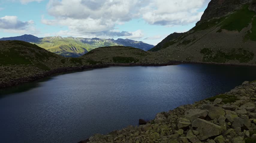 An aerial view moves forward over a high-altitude glacial lake. The water ripples gently amidst surrounding rocky terrain and mountains under scattered clouds.