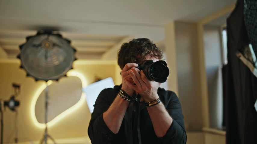 Male photographer directing photoshoot studio, gesturing hand signals giving instructions. Focused cameraman aiming DSLR camera model, professional creative shoot behind scenes work indoor space.