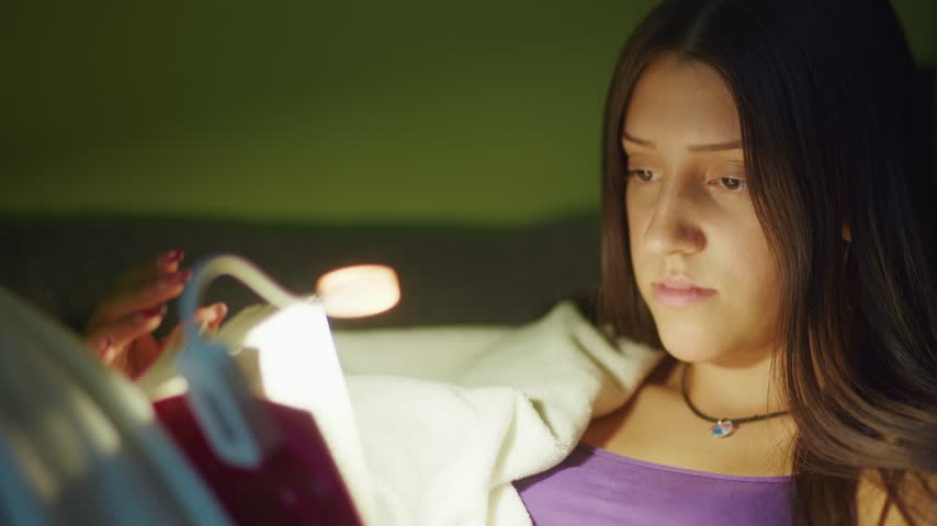 Cinematic close-up of a young girl reading a book at night, illuminated by a warm reading lamp. Cozy, intimate atmosphere for family, education and lifestyle projects