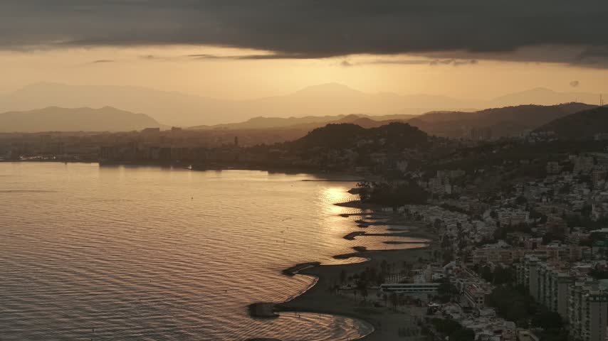 Aerial view west along the Malaga coast at sunset, with crescent breakwaters, beaches and apartment blocks leading toward the city and hills under a dark cloud.