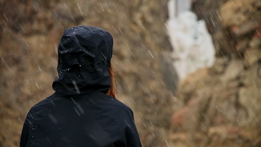 A woman stands facing away from the camera in a snowy outdoor environment. Snow falls around her in slow motion. The shot is a medium close-up from behind.