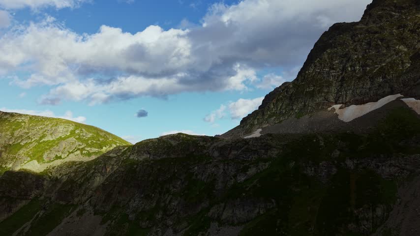 Clouds drift across the sky above rocky mountain peaks and grassy alpine slopes. The elevated wide shot shows high-altitude terrain with residual snow patches. Natural daylight illuminates the landscape.