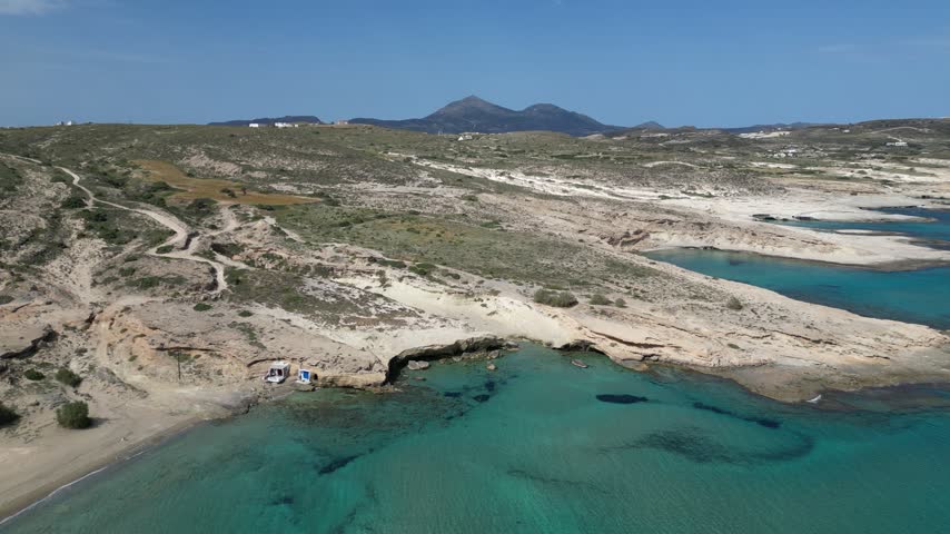 Drone footage of arid Greek island terrain with winding dirt trails leading to a turquoise cove and boats near the shore.