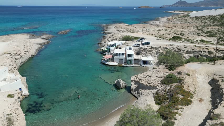 Drone shot of a small whitewashed fishing village on a rocky cove with clear turquoise water, boat houses, and people swimming near the shore.