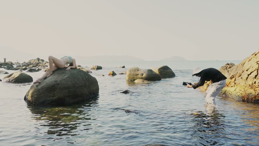 Backstage of bikini photo session with pretty female model and male photographer taking pictures with professional camera while lady is posing sitting on big stone on beach. Behind the scene of shoot.