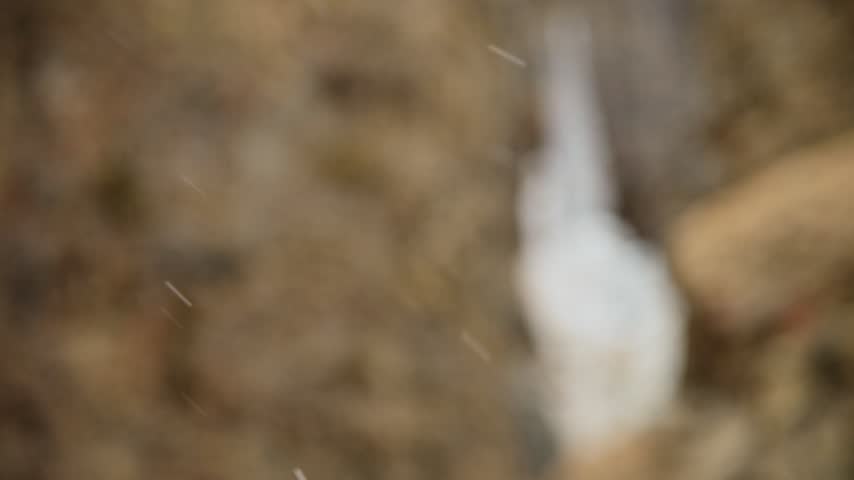 Snowflakes fly sideways in slow motion and shallow depth of field. Against the background of a frozen waterfall and a grassy dry slope