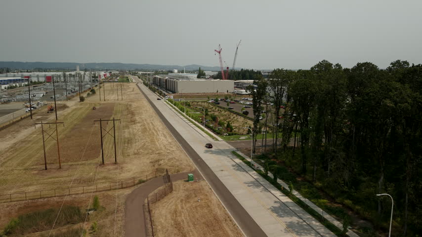 Cars driving along a road beside large data center buildings in Hillsboro, Oregon, with rooftop construction activity and a crane supporting technology expansion.