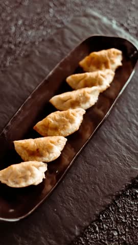 Close-up footage showcasing golden-brown dumplings artfully presented on a sleek, black platter. The dish sits atop a dark, textured slate background, creating a dramatic contrast. Suitable for culinary blogs, restaurant menus, and food-related promotional materials.