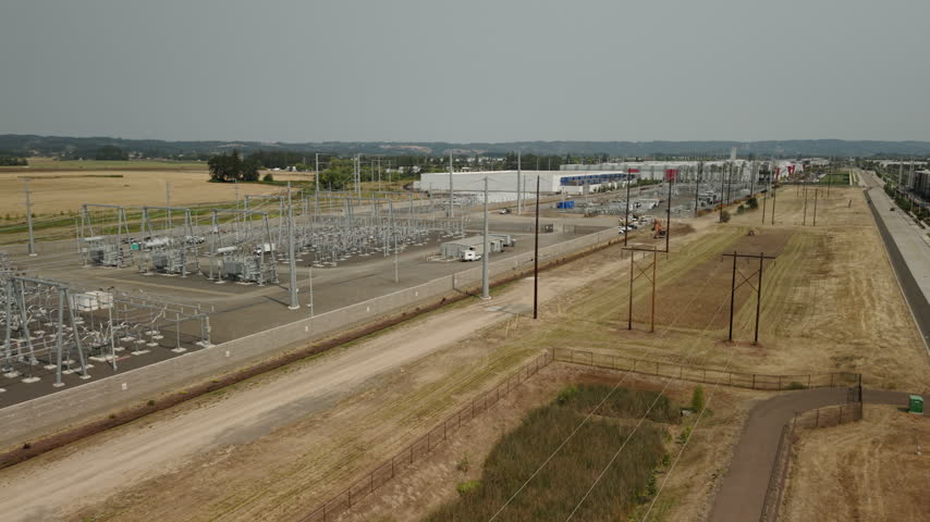 Aerial view of an electrical substation in Hillsboro, Oregon, showing transformers, transmission lines, and switching equipment near a major data center complex. Drone forwards flight.