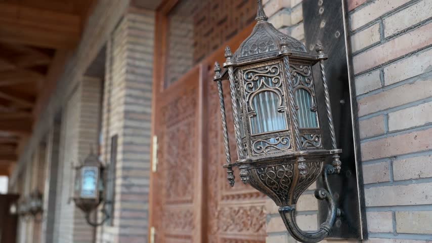 Close up view of ornate metal wall lantern beside a carved wooden door and patterned brick facade. Traditional Uzbek craftsmanship and central asian architectural detail.