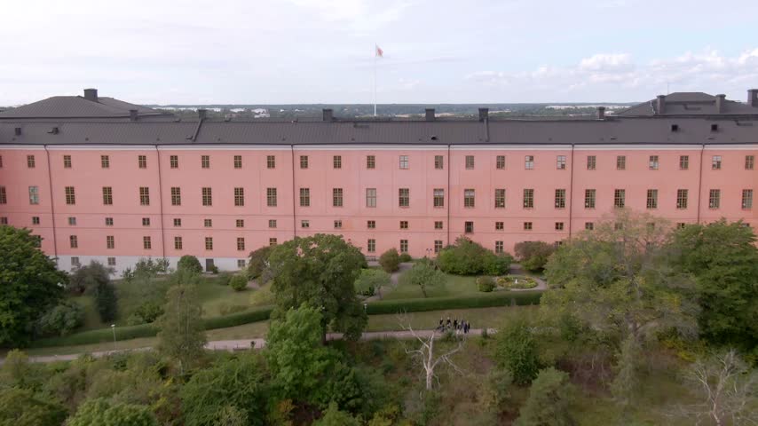Drone along salmon-pink Uppsala Castle facade and dark roof looking over trees toward the city horizon.