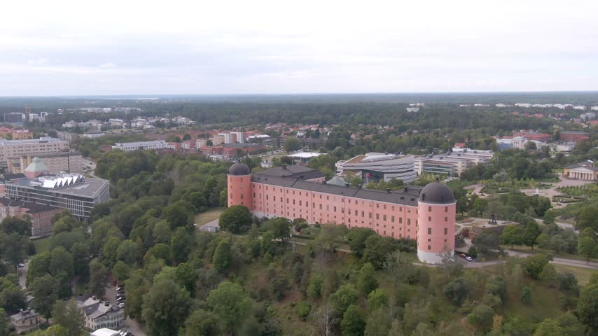 Diagonal aerial of pink Uppsala Castle on a wooded hill showing both black-domed towers and white buildings beyond.