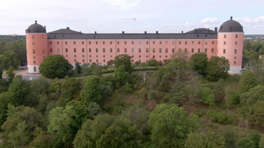 Wide aerial of pink Uppsala Castle on a wooded ridge with both black-domed cylindrical towers.