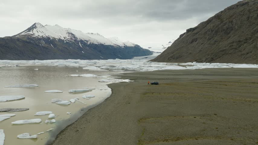 SUV approaching Fjallsarlon across the glacial outwash plain of Breidamerkursandur, with floating ice, Fjallsjokull and surrounding mountain slopes in South Iceland, drone forward flyover