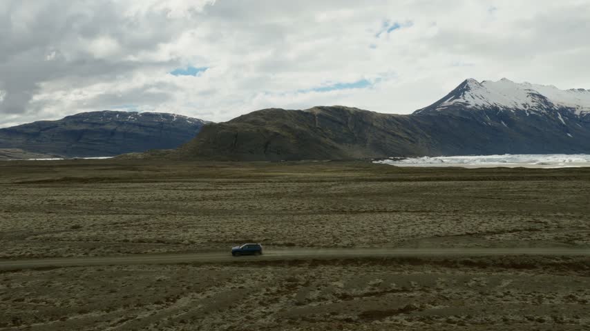 SUV driving fast on a dusty gravel track across the glacial outwash plain near Fjallsarlon, with Breidamerkurfjall and Vatnajokull terrain rising behind, South Iceland, drone tracking