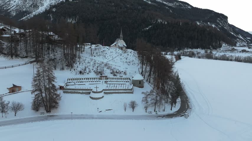 Aerial view of a historic church on a hill surrounded by a snow-covered cemetery and winter landscape in Graun im Vinschgau, Trentino-Alto AdigeS, Italy.