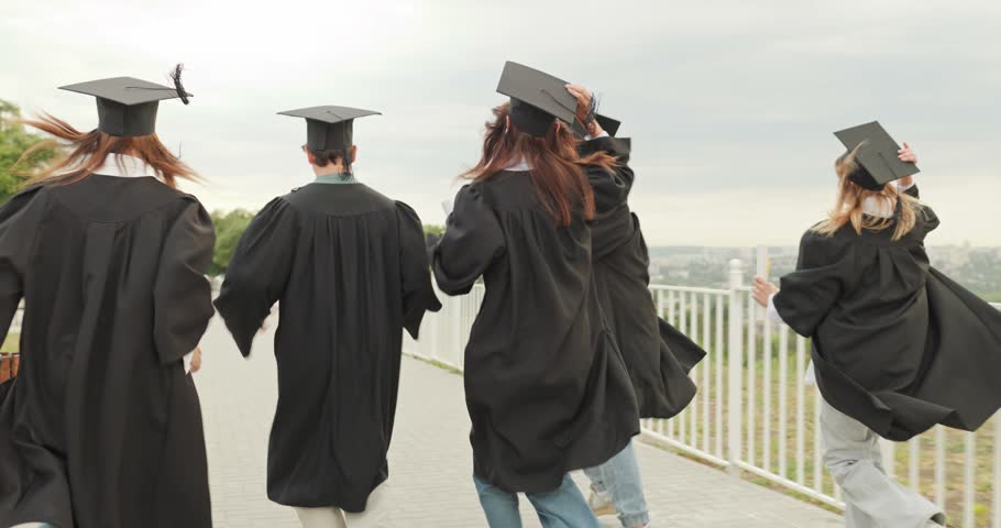 Graduates celebrate graduation while running and jumping outdoors. Footage follows friends in gowns and caps along a park walkway, holding diplomas and raising arms. Pure joy marks a milestone.