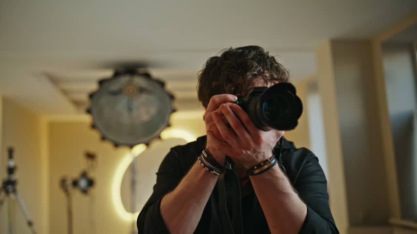 Photographer holding professional camera pointing lens at viewer, taking shots indoors studio. Cheerful young man gesturing stop, laughing during creative photoshoot session, handheld shot studio.