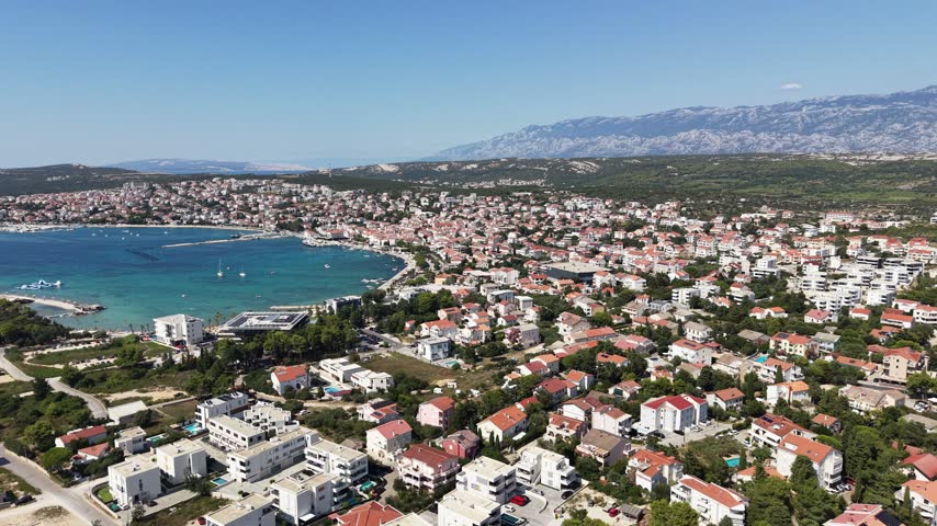 Aerial panorama of a coastal town beside a blue bay in Croatia, with red roofs, a waterfront and distant mountains in sunshine.