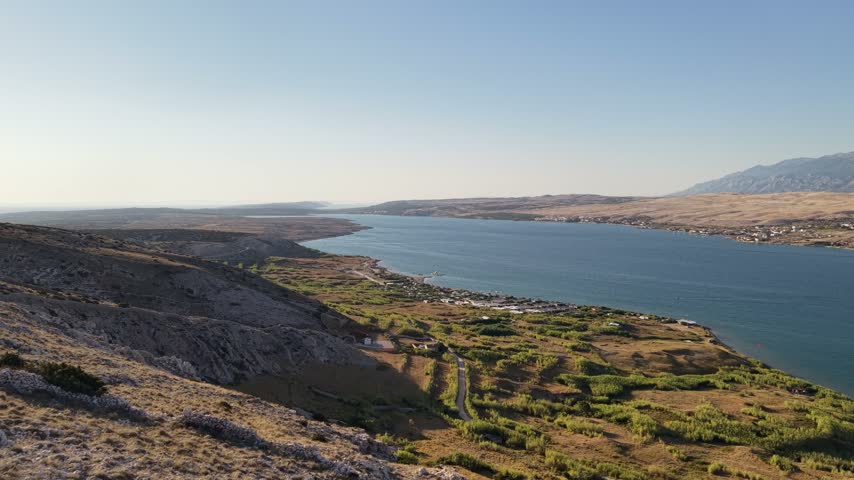 Aerial view of a green coastal slope beside a blue bay in Croatia, with rocky hills and distant mountains in warm sunlight.
