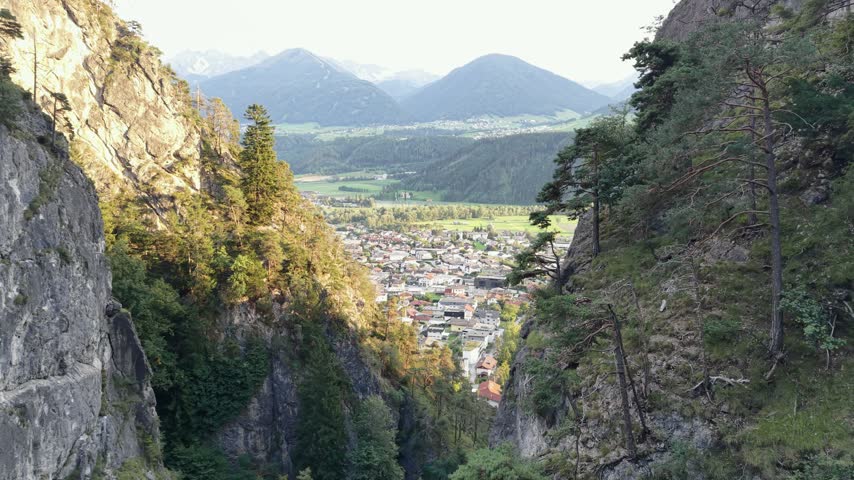 Drone reveal between rocky cliffs toward a town and mountain valley in Tyrol, Austria, with trees and sunlit green fields.