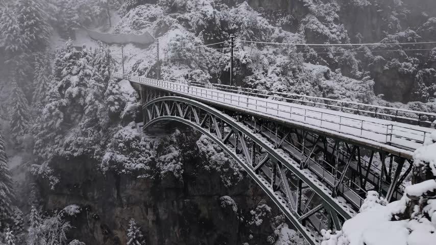 Drone view moving alongside a snow-covered railway arch bridge spanning a forested gorge in winter, Tyrol, Austria. Soft winter daylight.