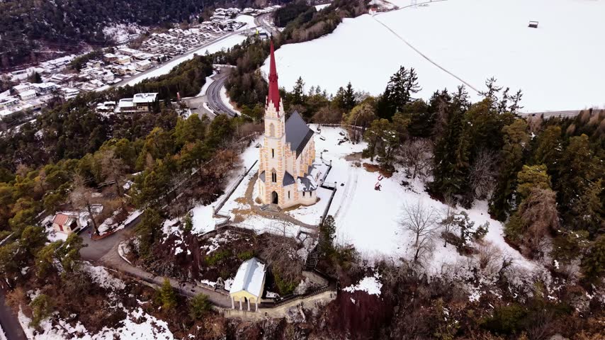 Drone orbit around a hilltop church in a snowy landscape in Tyrol Austria. Soft winter daylight.