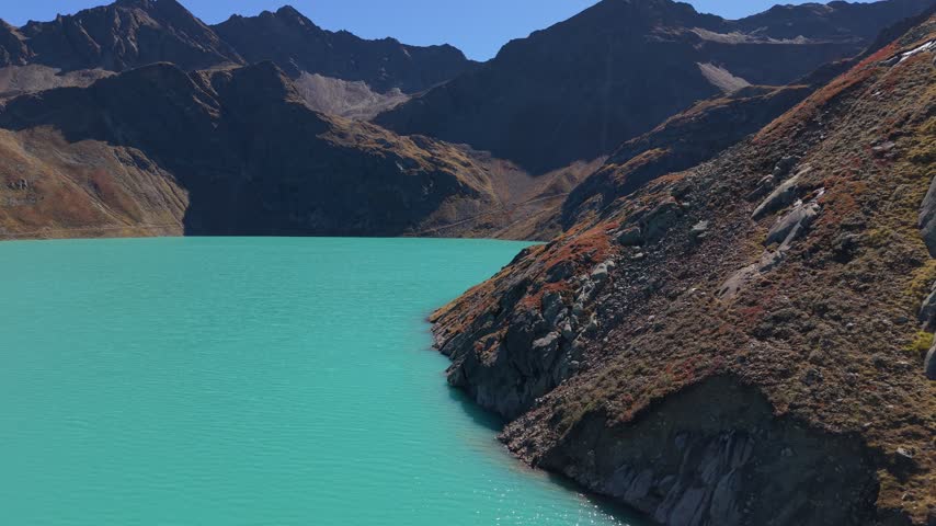A continuous curved aerial view follows a rocky projection into turquoise reservoir water, with reddish alpine vegetation, shaded mountains and clear sunny weather. Tyrol, Austria.