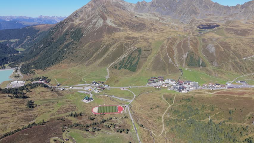 A continuous aerial panorama shows sunlit meadows, mountain buildings, a sports field and distant reservoir beneath brown alpine slopes and a clear sky. Tyrol, Austria.